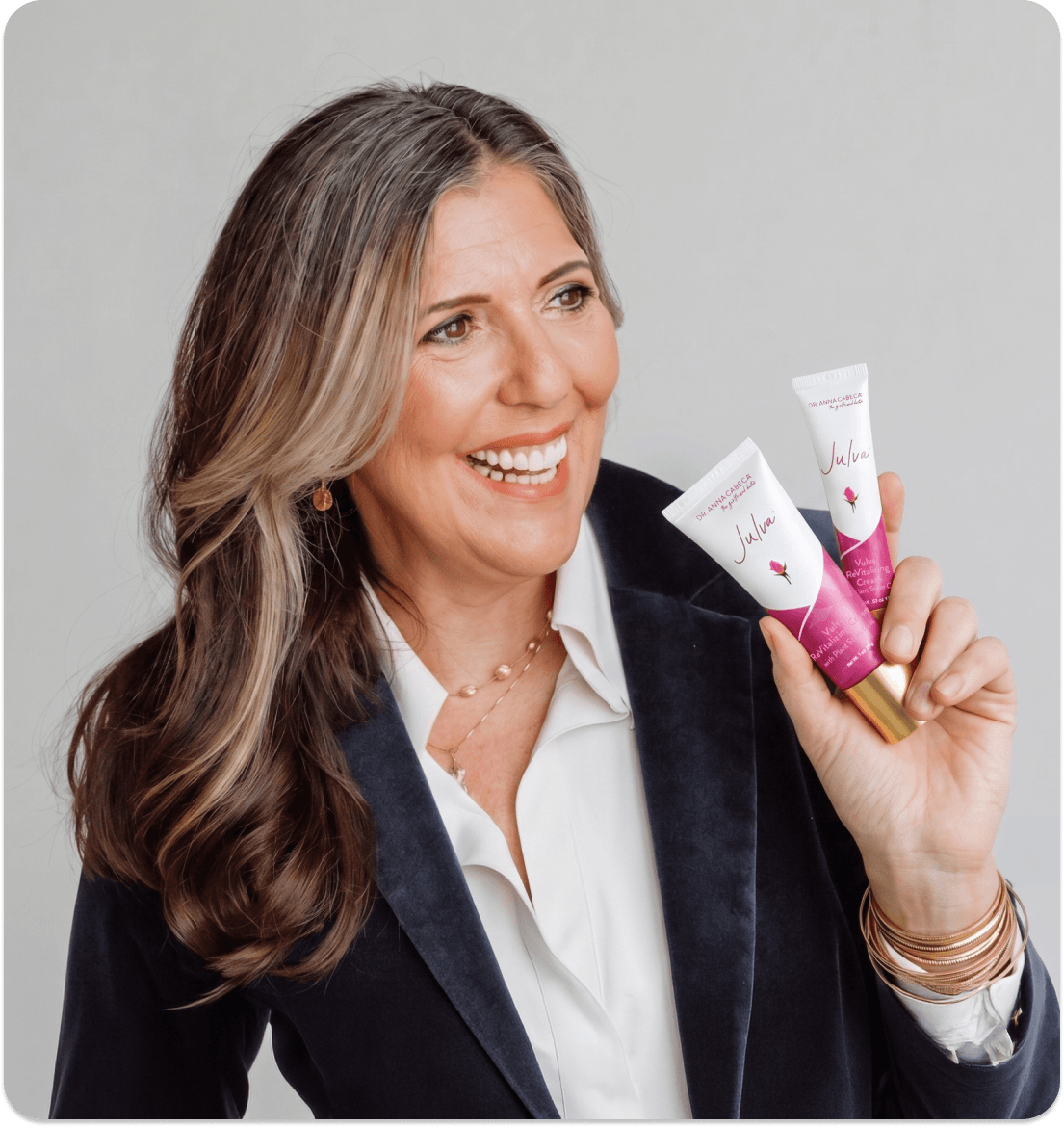Dr. Anna Cabeca holding two cosmetic tubes branded 'Julva' in a studio setting. She is wearing a white collared shirt and a dark blazer against a light gray background.
