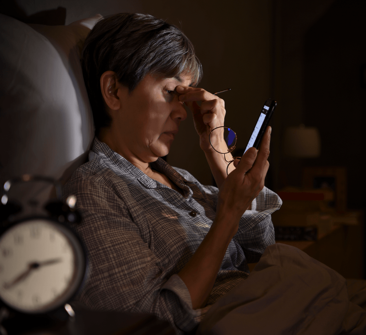 An older woman lying in bed at night looks at her smartphone, with a bedside alarm clock visible in the foreground and dim bedroom lighting around her.