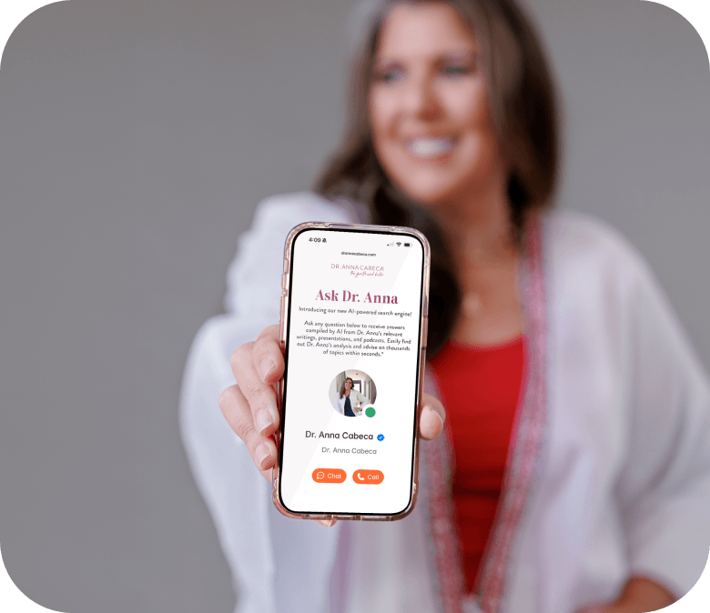 Dr. Anna Cabeca in a white blazer and red top holds up a smartphone showing a digital profile page titled 'Ask Dr. Anna' against a soft gray studio background.