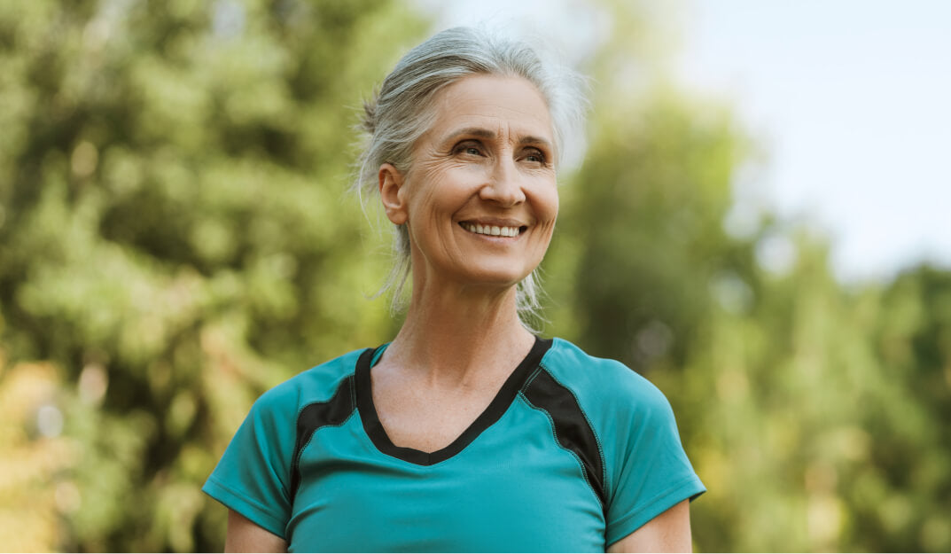 Woman with gray hair wearing a teal shirt outdoors with greenery in the background