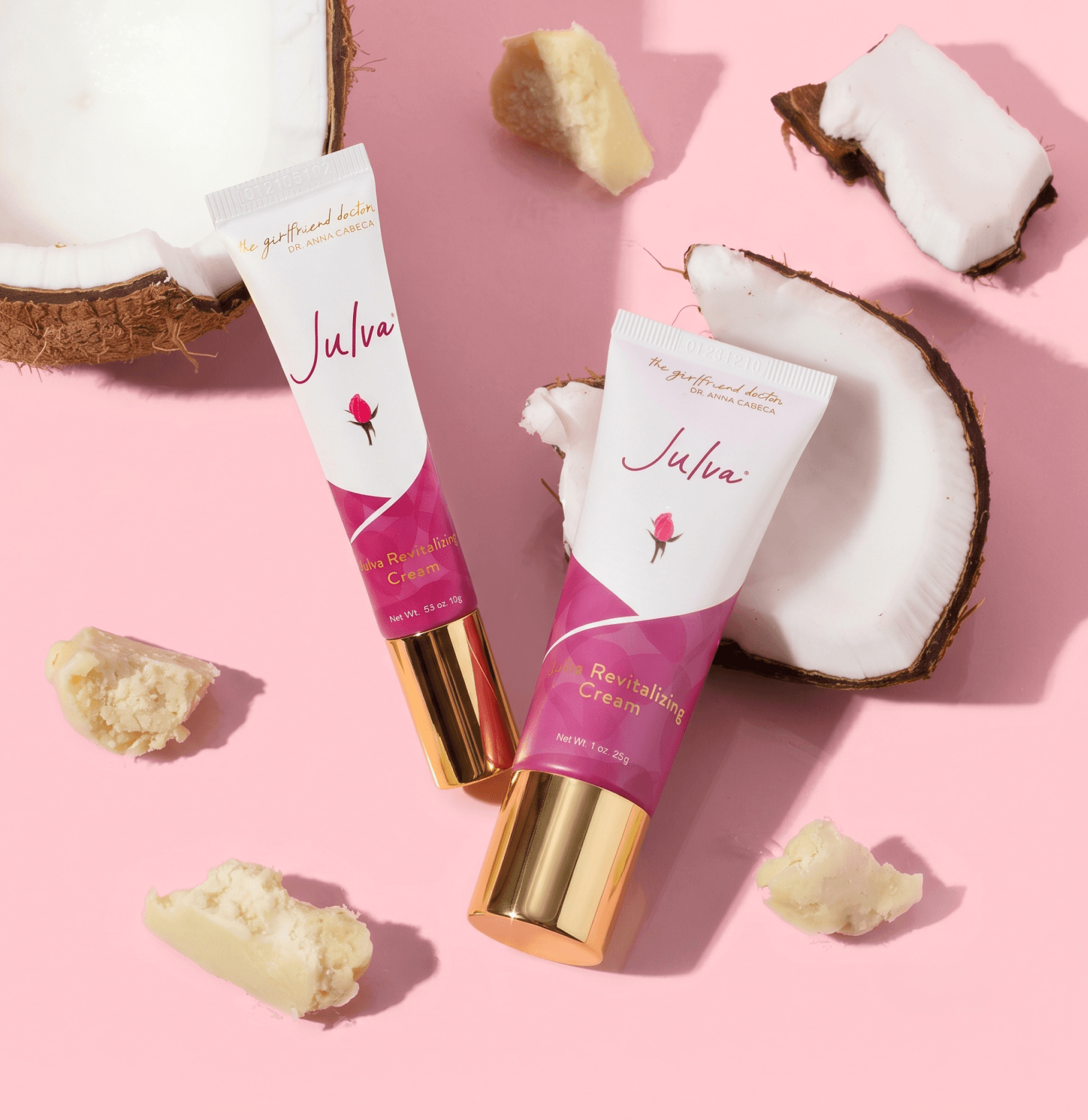 Two tubes of Julva Revitalising Cream are arranged on a pink background with coconut halves and cream pieces as styled props. The tubes are white with a magenta-pink label and metallic gold caps.