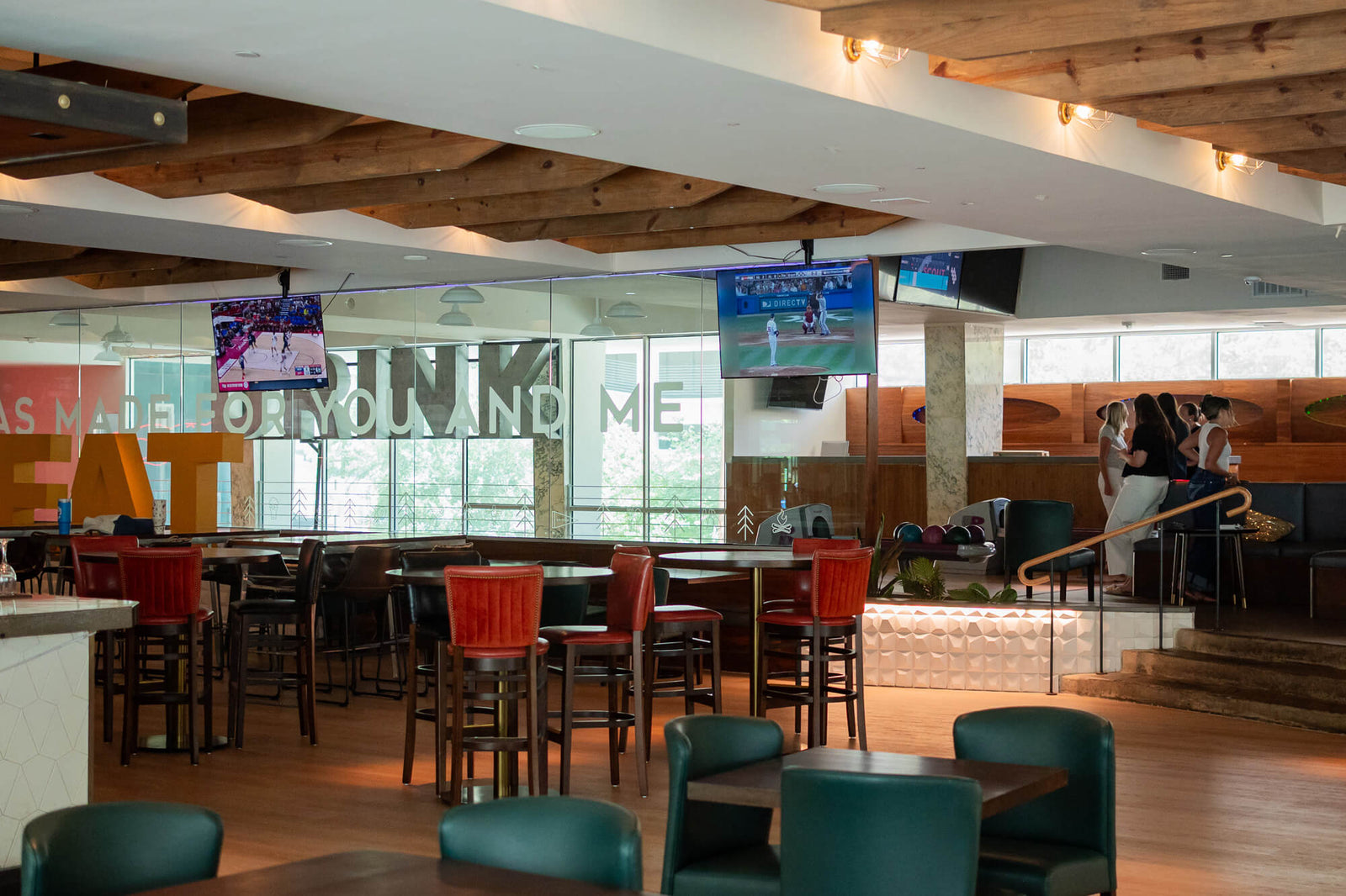 A large indoor restaurant or bar seating area with wooden ceiling beams, multiple hanging TVs, bar stools, tables, and glass walls. No single retail product is isolated as the subject.