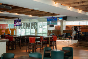 A large indoor restaurant or bar seating area with wooden ceiling beams, multiple hanging TVs, bar stools, tables, and glass walls. No single retail product is isolated as the subject.