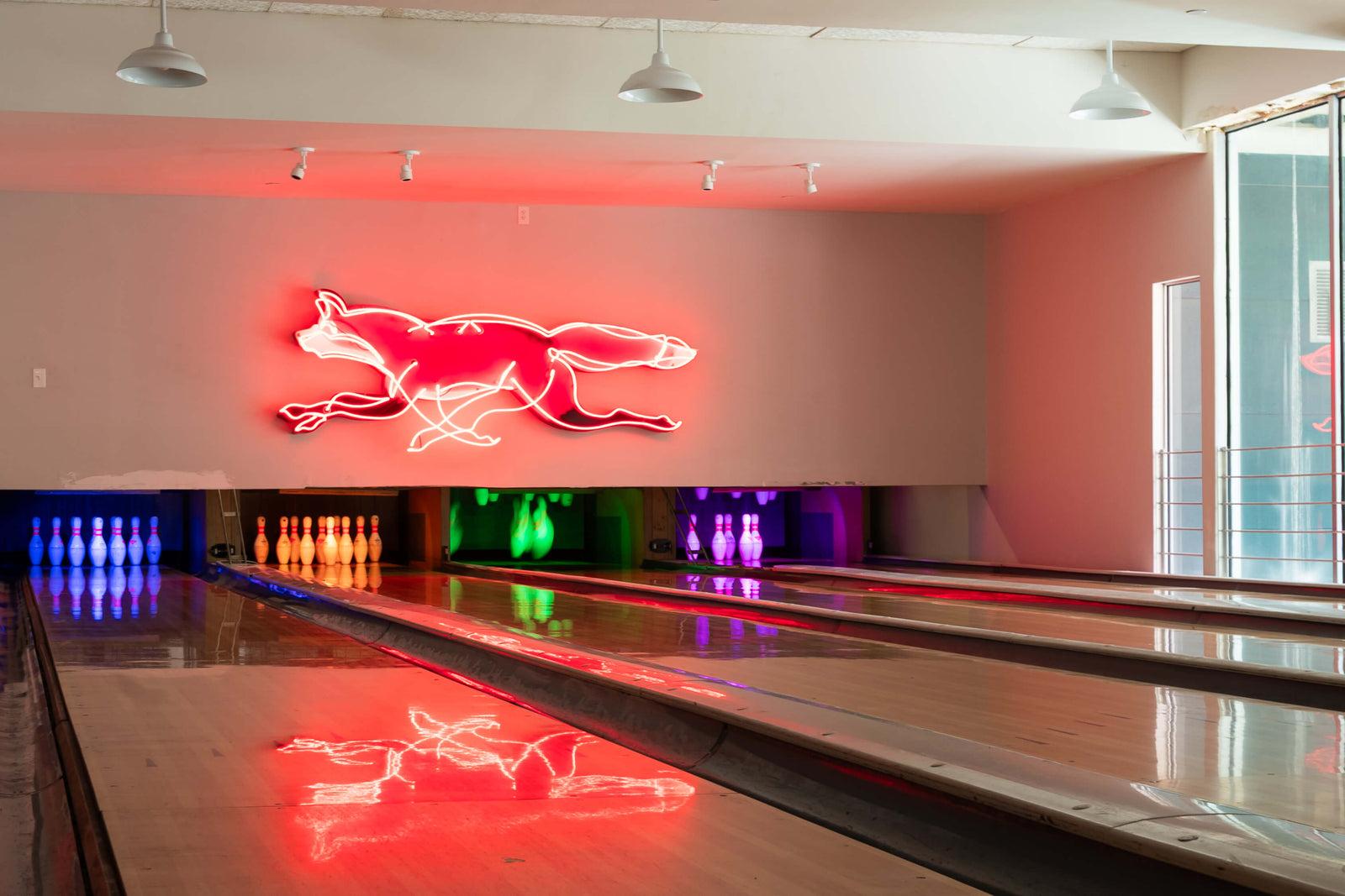A neon sign of a running fox in red and white glowing on the wall of a bowling alley, with bowling lanes and colorful pin lights visible below.
