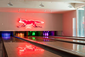 A neon sign of a running fox in red and white glowing on the wall of a bowling alley, with bowling lanes and colorful pin lights visible below.