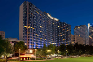 The Stater hotel at dusk, shown as a tall illuminated skyscraper with blue and yellow lights against a deep blue evening sky. Trees, a lit PARK sign, and a grassy foreground are visible in the lower portion of the frame.